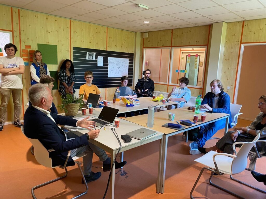 A group of people sit and stand in a meeting room with wooden walls and an orange floor. About 10 people sit at the table, while a few stand at the back of the room. On the table are laptops, cups, snacks and bottles of drinks. One person, the facilitator, talks while others listen intently. A bulletin board with drawings and notes is visible in the background.     